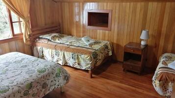 Quarto com duas camas, paredes de madeira, uma pequena mesa de cabeceira com abajur e uma janela com cortinas.