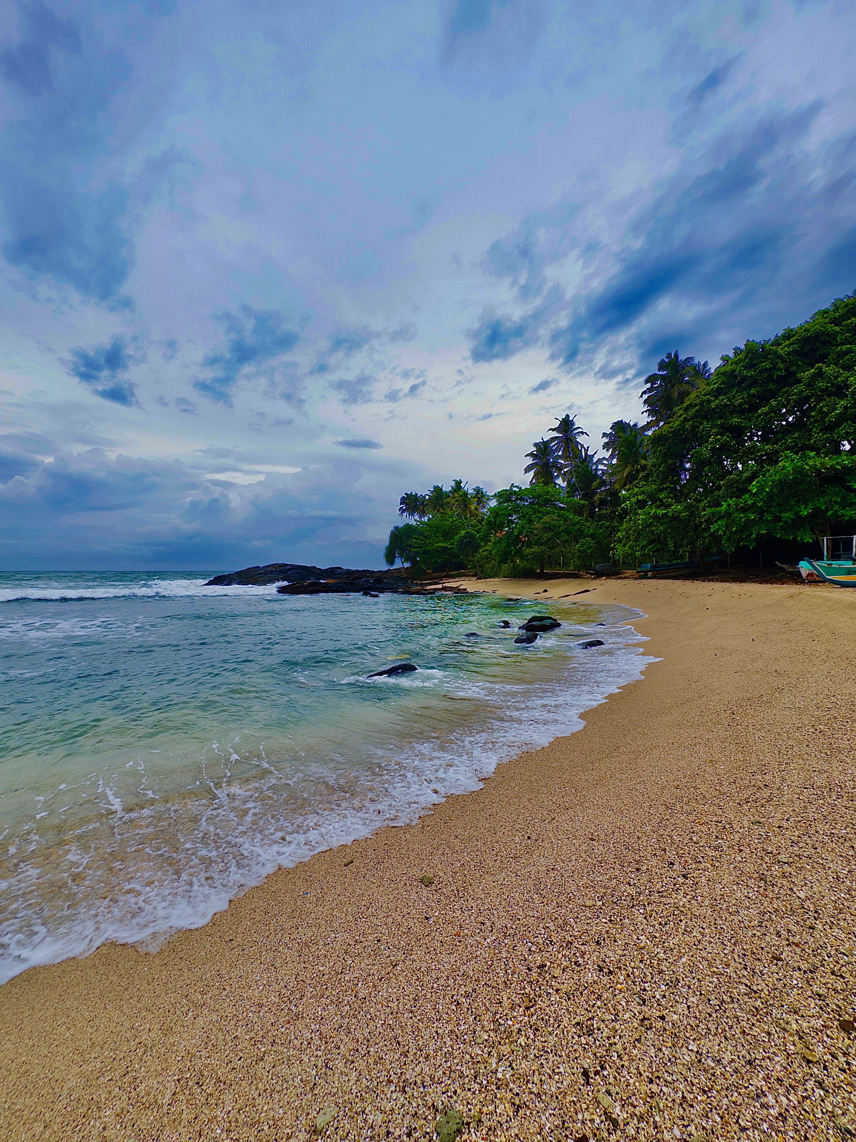 A beach with pebbly shore, calm sea, and dense foliage in the background.