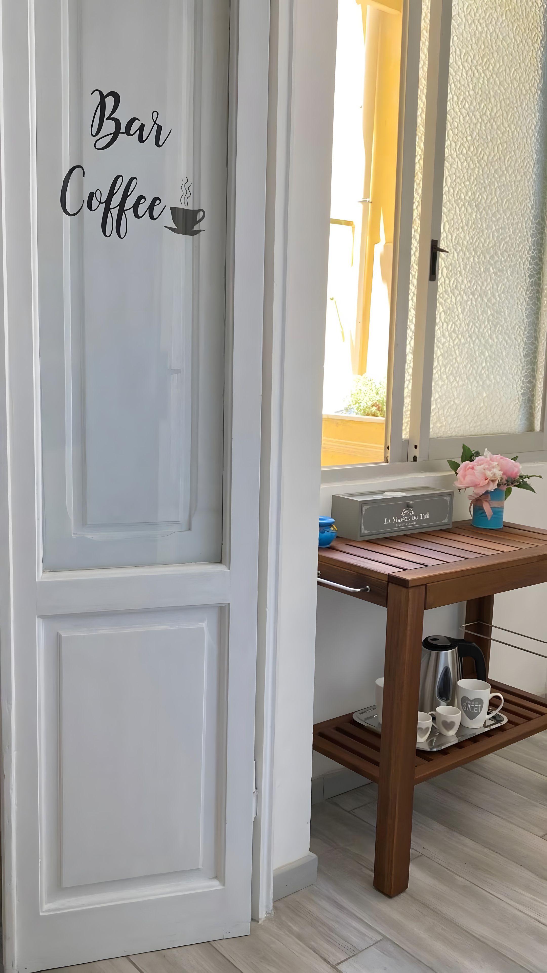 Een witte deur met 'Bar Coffee' aanduiding, een houten tafel met een waterkoker, een dienblad en een mok met de tekst 'sweet', en een raam met een bloevas.