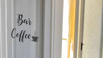 Een witte deur met 'Bar Coffee' aanduiding, een houten tafel met een waterkoker, een dienblad en een mok met de tekst 'sweet', en een raam met een bloevas.