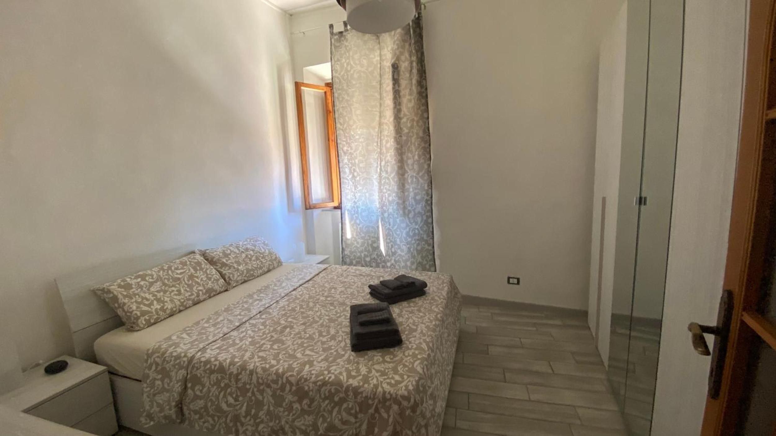 Een kleine, goed verlichte slaapkamer met een bed, een nachtkastje en een raam met gordijnen.