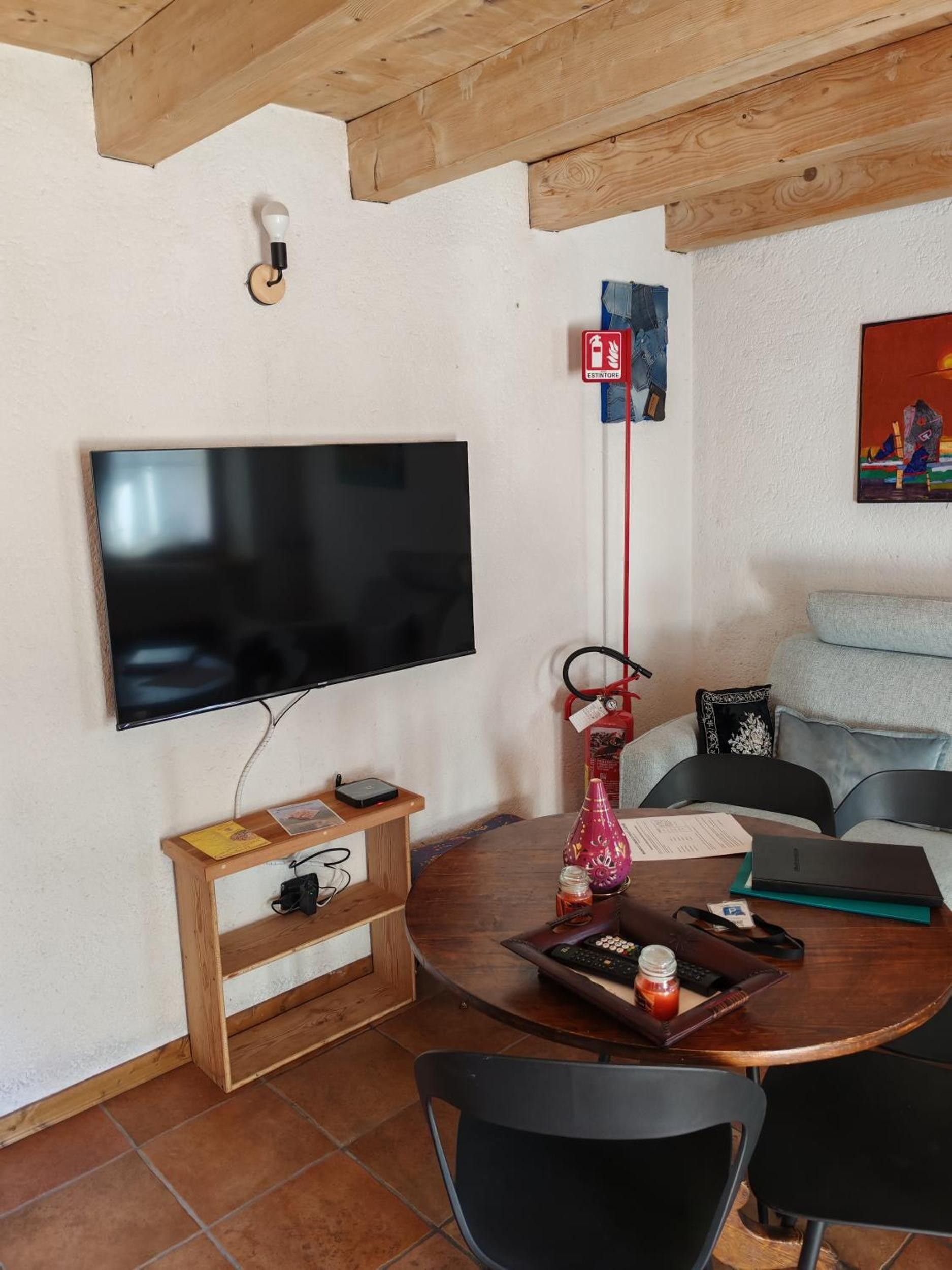 A living room with a flat-screen TV mounted on the wall, a wooden table with a vase, a fire extinguisher, and a sofa.