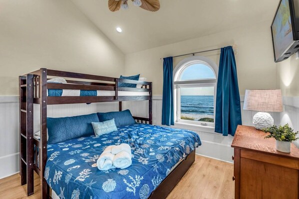Une chambre avec un lit superposé, une grande fenêtre avec des rideaux, une table de chevet avec une lampe, et une vue sur la mer.