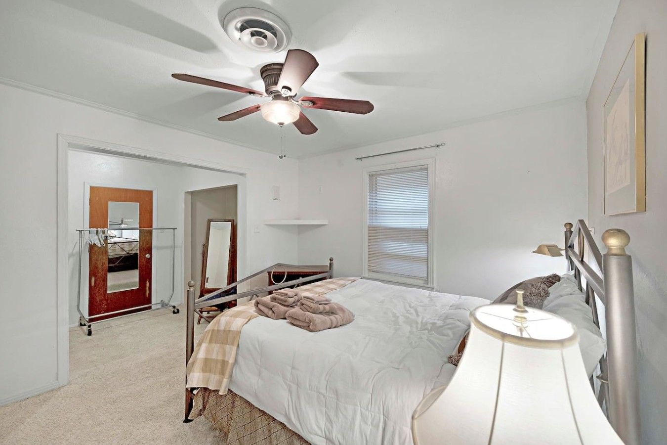 A bedroom with a bed, a ceiling fan, a bedside lamp, a mirror, and a window with blinds.