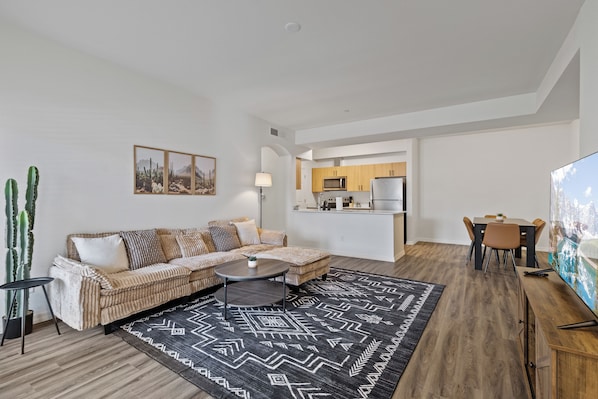 A modern living room with a sofa, coffee table, and a dining area with chairs and a TV. - Modern Power Ranch Retreat - Pool & Spa (Gilbert)