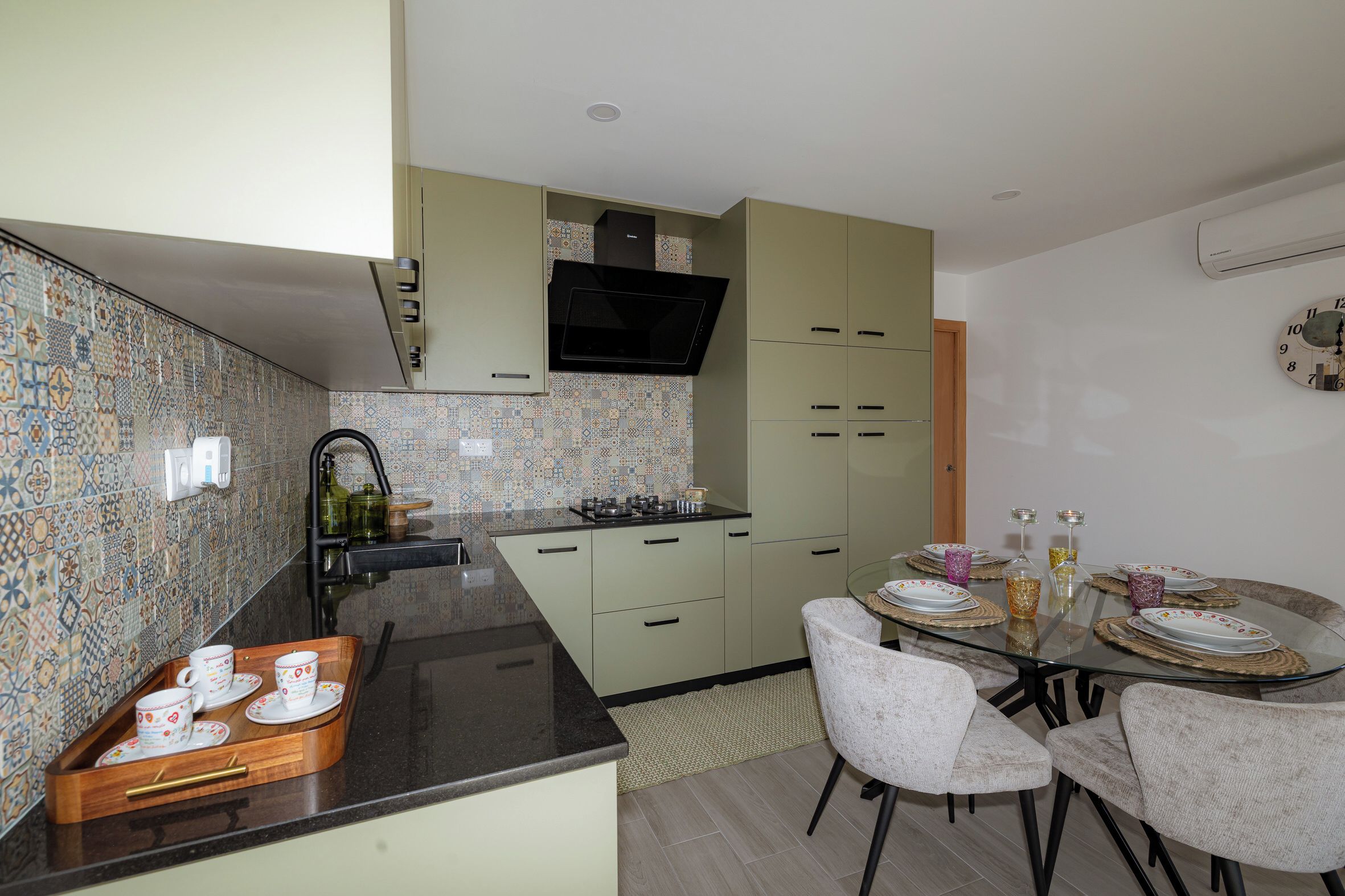 Una cucina moderna con zona pranzo, caratterizzata da un piano di lavoro nero, mobili chiari color verde e una parete di fondo rivestita di piastrelle decorate.