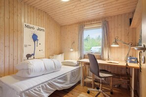 Ein Zimmer mit einem Bett, einem Schreibtisch und einem Sessel. Es gibt ein Fenster mit Vorhängen, ein Poster an der Wand und eine Lampe auf dem Schreibtisch.