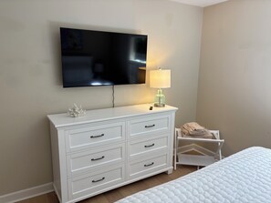 A bedroom with a white bed, a white dresser with drawers, a flat-screen TV mounted on the wall, and a bedside table with a lamp. - New Listing!  Beautifully Furnished 3 Bedroom in Sea Pines (Hilton Head Island)
