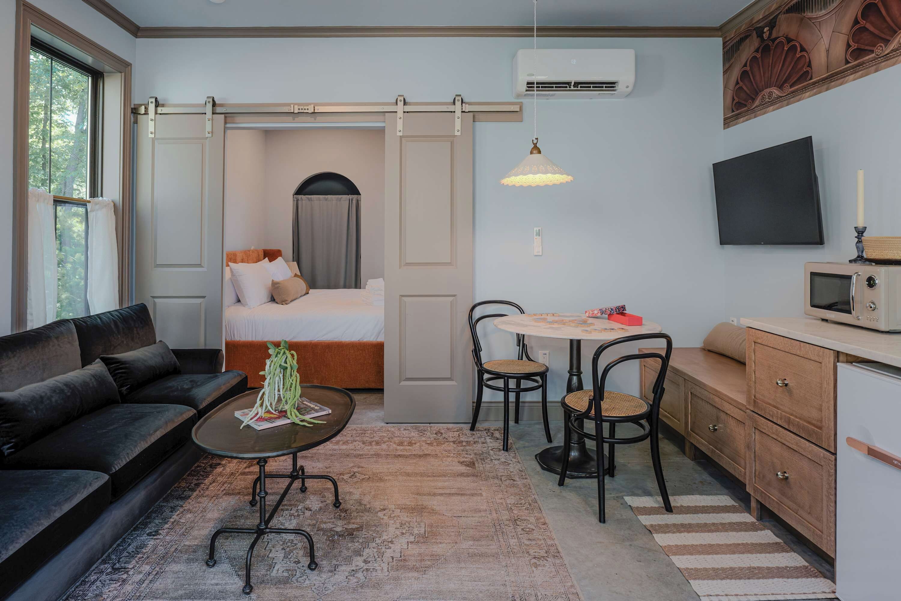 A compact hotel room with a sofa, a bed, a dining area with chairs, a small table, a microwave, and a TV on the wall.