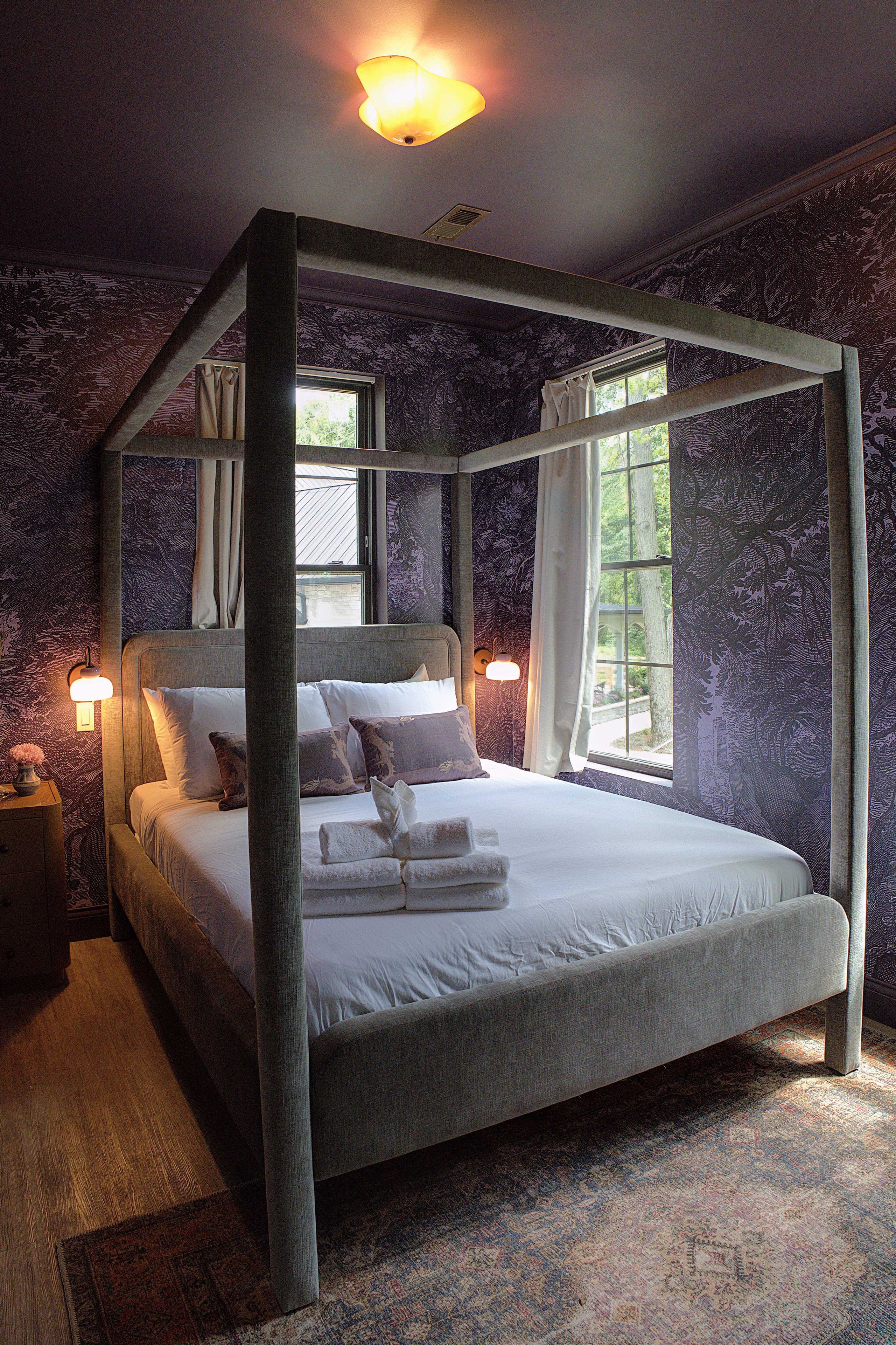 A four-poster bed with a canopy, a nightstand, a lamp, and a window with curtains.
