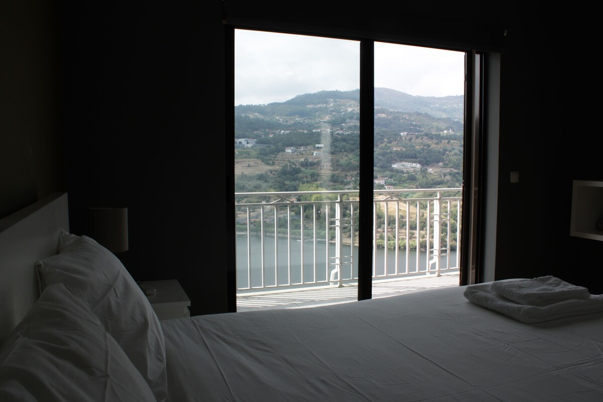 A hotel room with a large bed, a nightstand, a lamp, and a view of a balcony with a railing and a body of water.