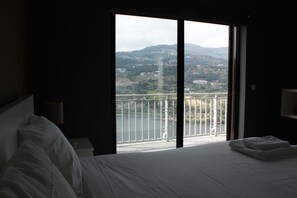 A hotel room with a large bed, a nightstand, a lamp, and a view of a balcony with a railing and a body of water. - Douro Nest House -  (Resende)