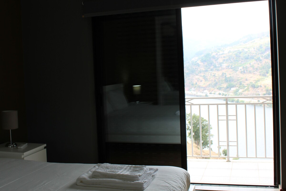 A bedroom with a bed, a nightstand, a lamp, and a view of a landscape through a large window.