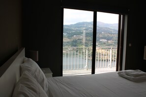 A hotel room with a large bed, a nightstand, a lamp, and a view of a balcony overlooking a body of water and a hilly landscape. - Douro Nest House -  (Resende)