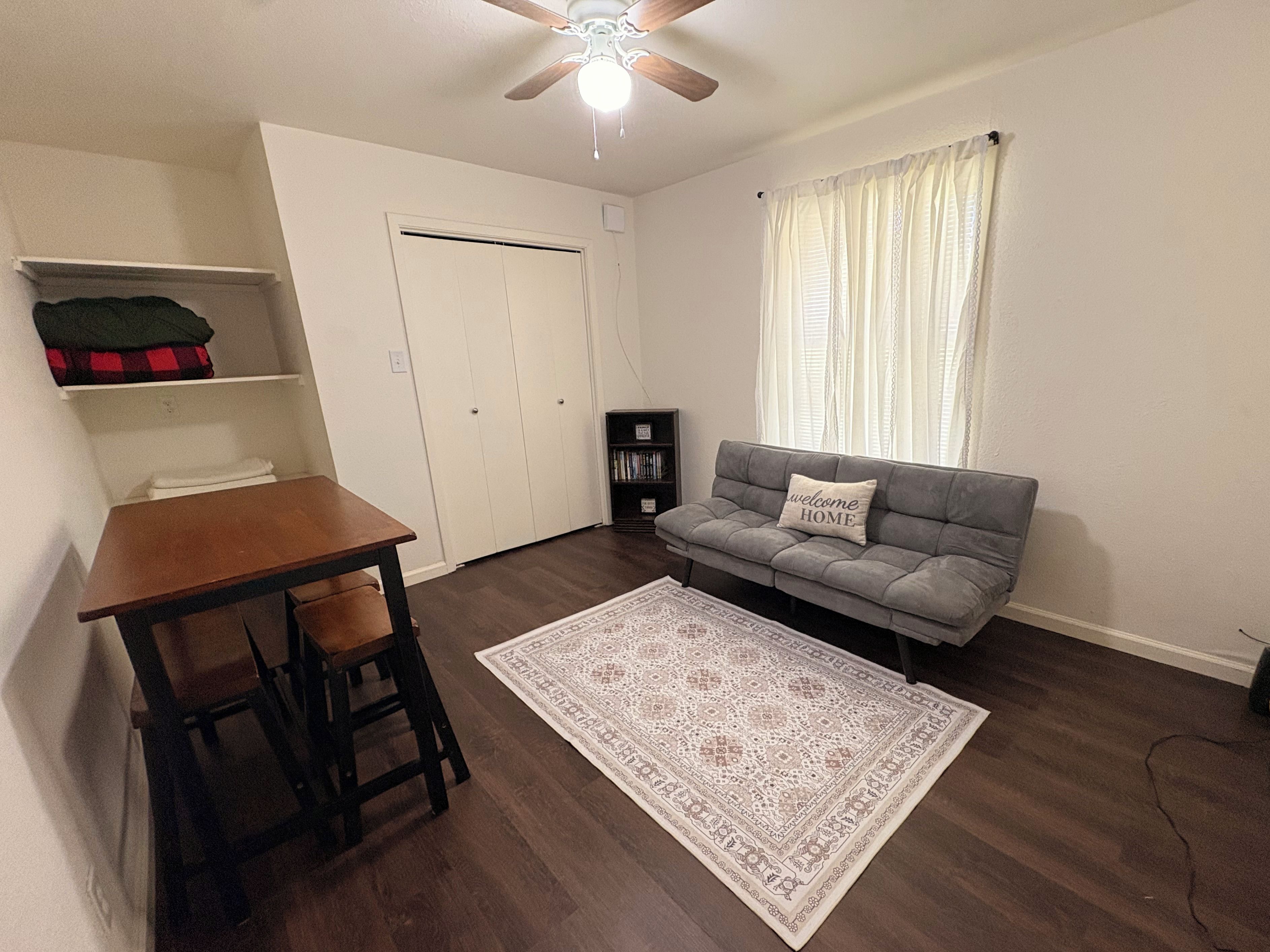 A room with a wooden dining table, a grey sofa, a patterned rug, a ceiling fan, and a window with curtains.