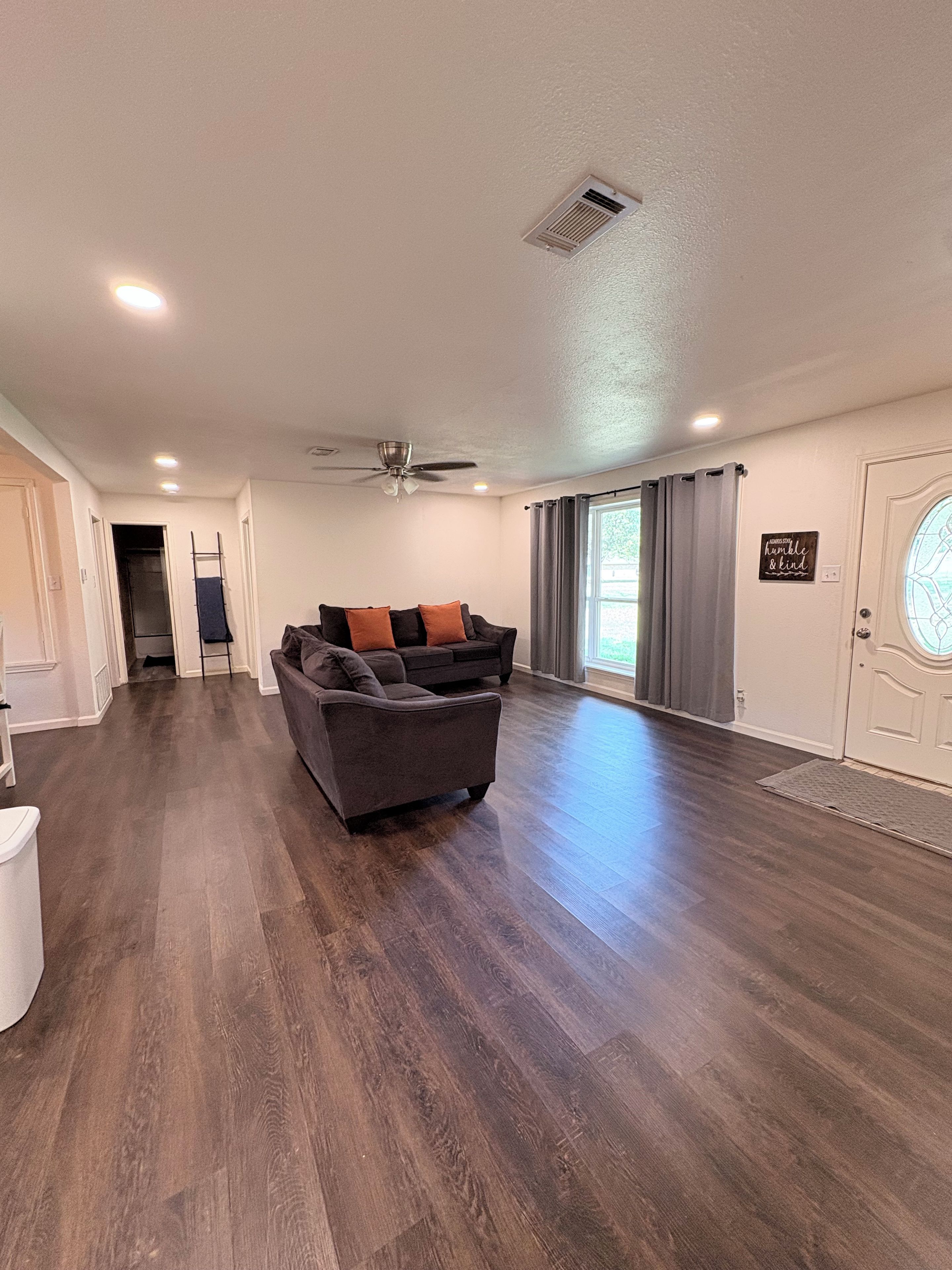 A spacious living room with hardwood flooring, a gray sofa with orange cushions, a ceiling fan, and a door with a frosted glass window.