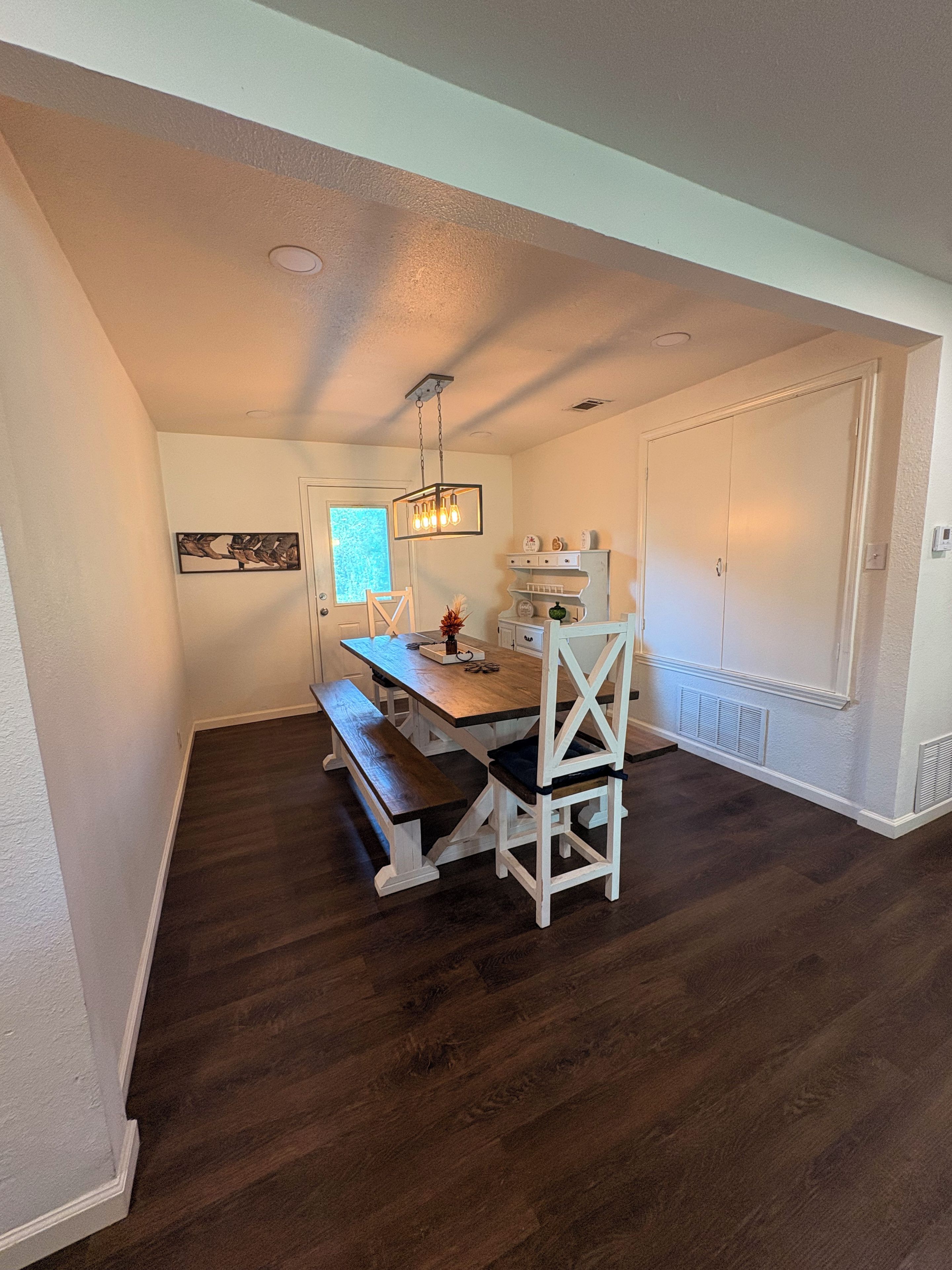 A dining area with a wooden table, white chairs, a bench, and a hanging light fixture.