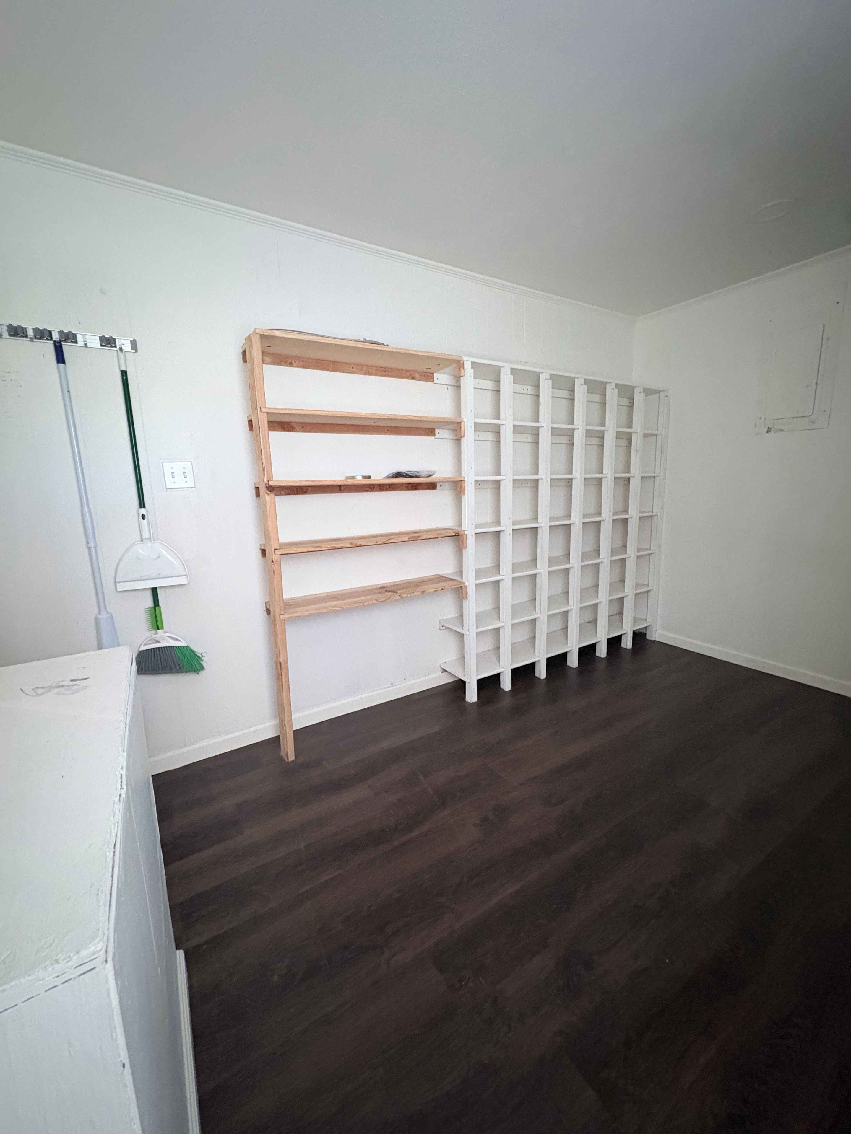 A room with a dark wooden floor, white walls, and a white shelving unit against the wall.