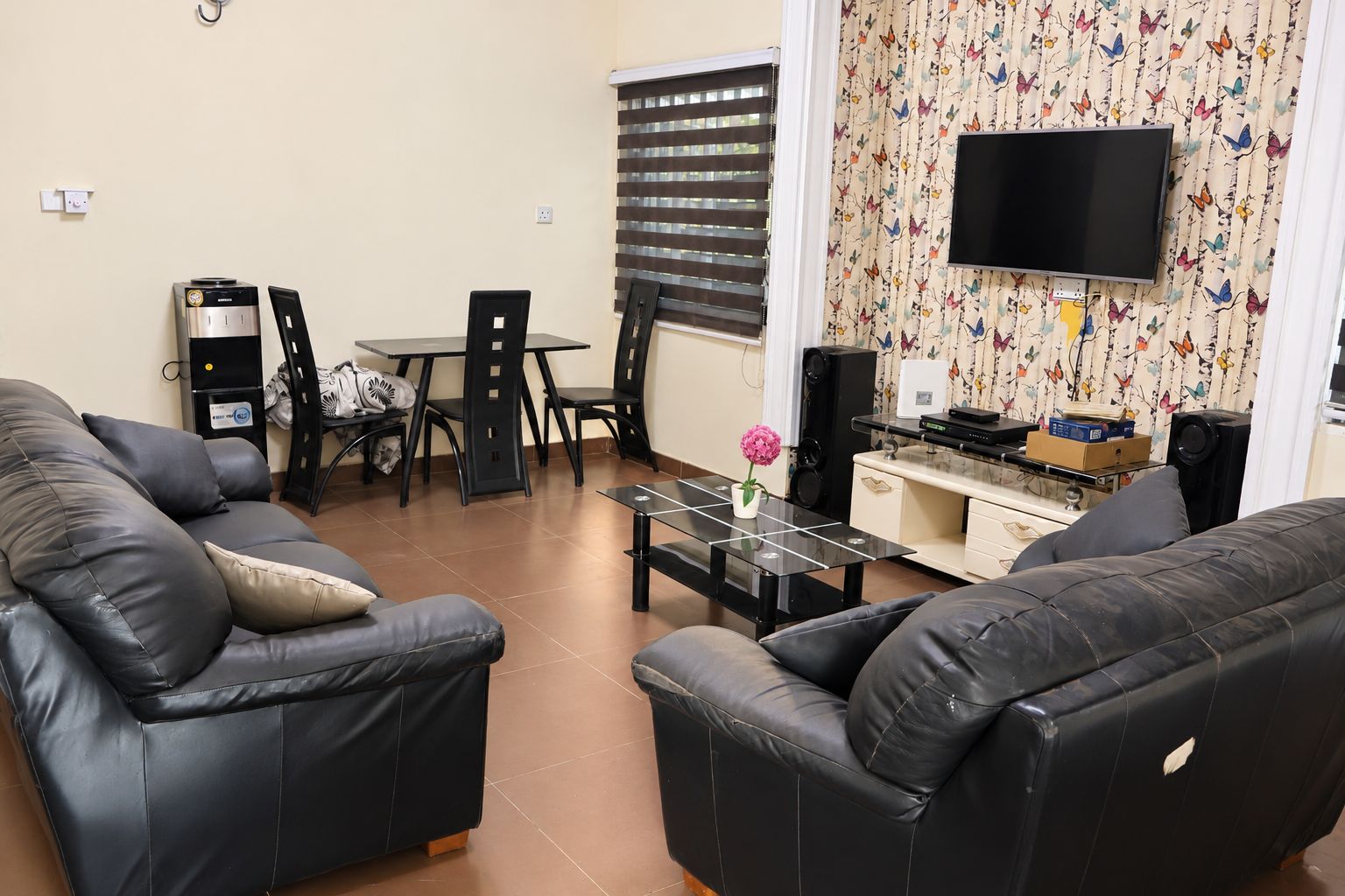 A living room with two black leather sofas, a glass coffee table, a dining table with chairs, a TV mounted on the wall, and a patterned curtain.