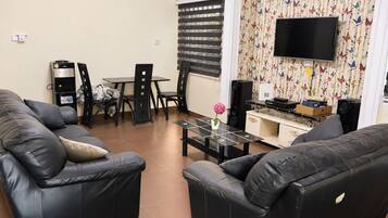 A living room with two black leather sofas, a glass coffee table, a dining table with chairs, a TV mounted on the wall, and a patterned curtain.