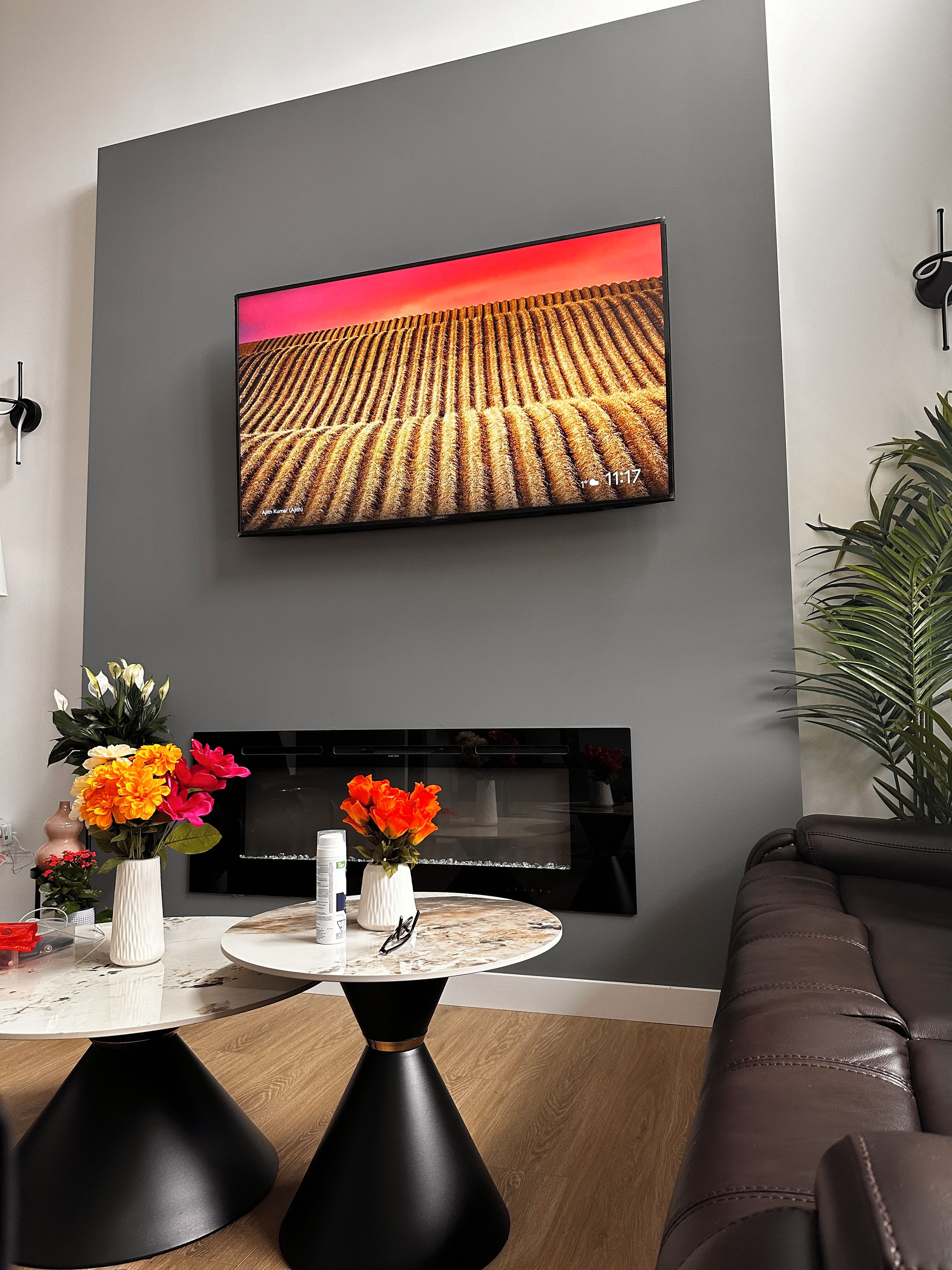 Een moderne woonkamer met een flatscreen televisie aan de muur, een haard eronder en een ronde salontafel met een bloemenvaas.