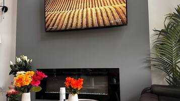 Een moderne woonkamer met een flatscreen televisie aan de muur, een haard eronder en een ronde salontafel met een bloemenvaas.