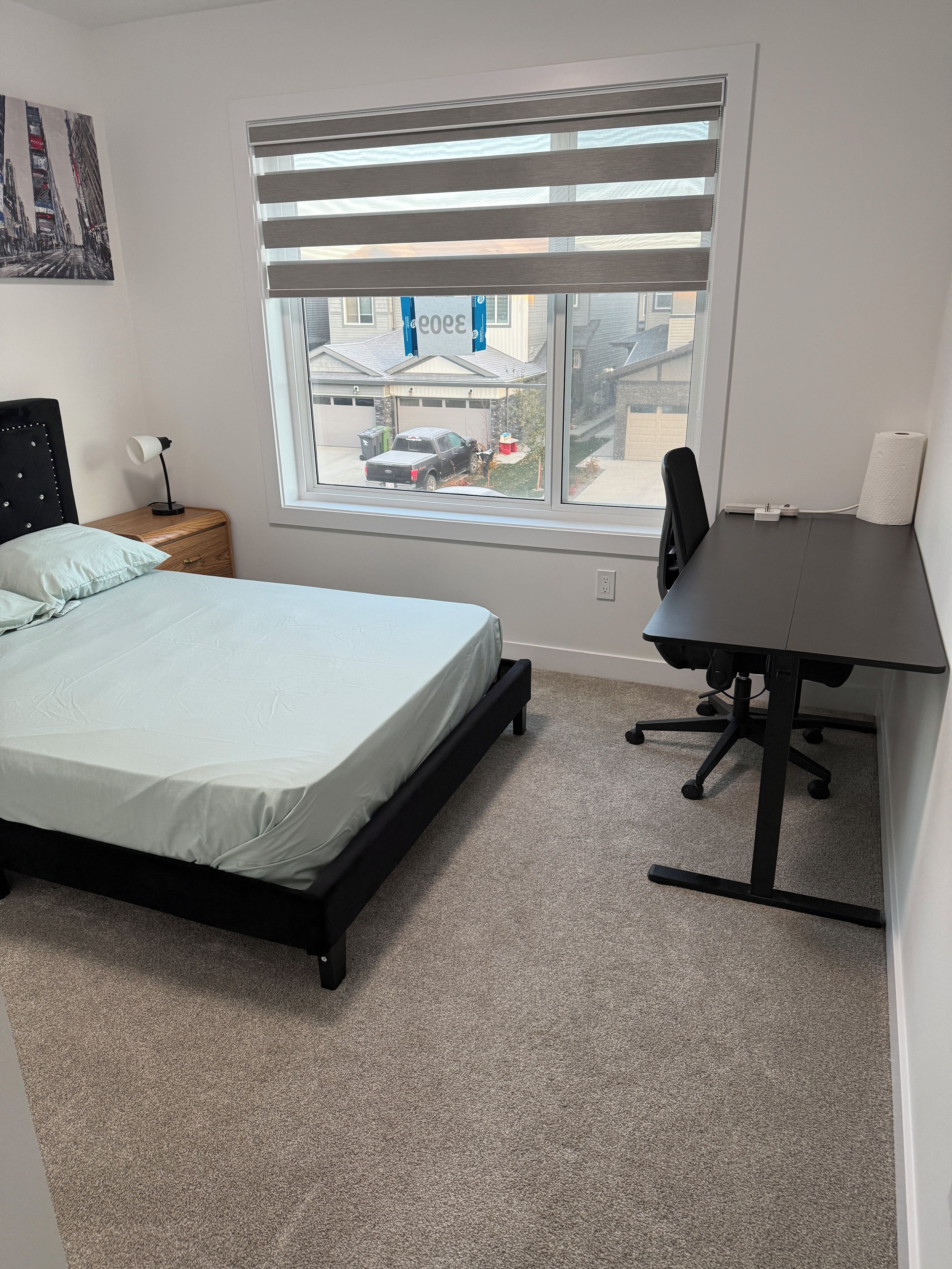 Een slaapkamer met een bed, een bureau, een stoel, een nachtkastje, een raam met jaloezieën en een schilderij aan de muur.