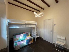 Een stapelbed met een kleurrijk matras, een witte stoel en een deur in een kamer met donkere betegelde vloer en houten balken aan het plafond.