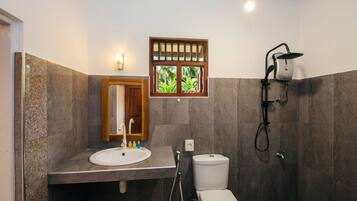 Un baño con lavamanos blanco, inodoro, ducha con cabezal de ducha manual y ventana con vistas a zonas verdes.