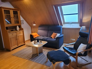 A cozy loft bedroom with a sloped ceiling, a large window, a wooden cabinet, a sofa, a chair, and a small table.