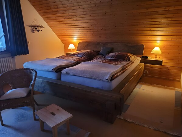 A cozy bedroom with a wooden ceiling, a bed with a plaid blanket, two bedside lamps, and a wicker chair.