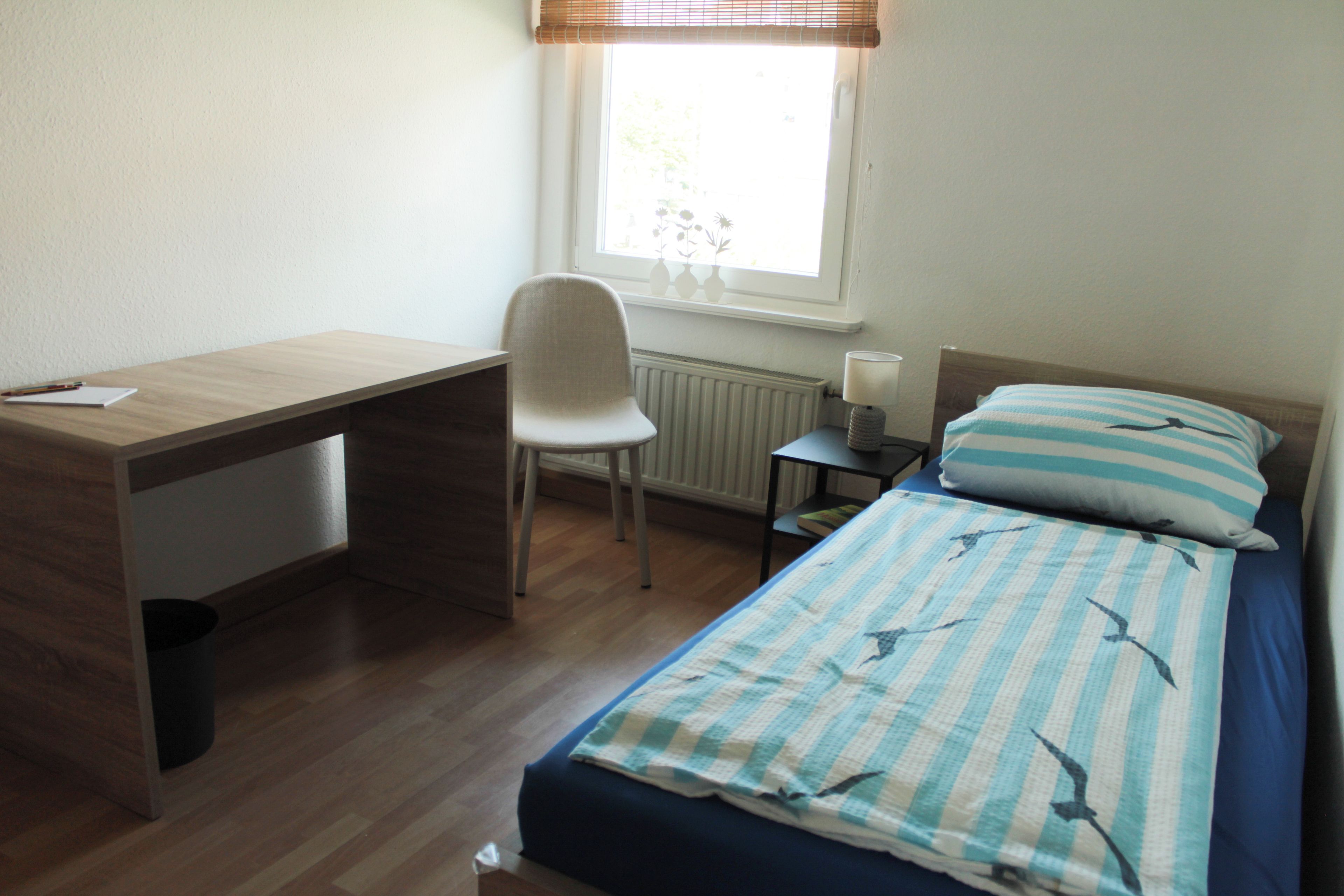 A bedroom with a wooden desk, a chair, a bedside table with a lamp, and a bed with striped bedding.