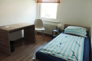 Quarto com escrivaninha de madeira, uma cadeira, mesa de cabeceira com abajur e cama com roupa de cama listrada.