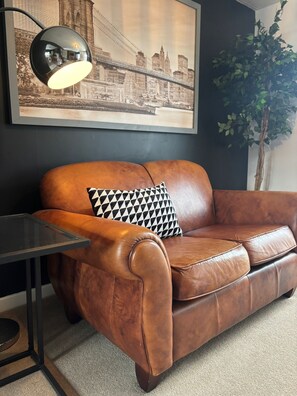 Un canapé en cuir marron agrémenté d’un coussin à motifs noir et blanc, un lampadaire noir et, au mur, un tableau représentant une vue urbaine encadré.