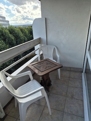 Ein Balkon mit einem weißen Plastikstuhl, einem kleinen Tisch und Blick auf ein Gebäude in der Ferne.