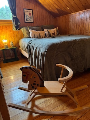 Ein gemütliches Schlafzimmer mit Holzbalkendecke, einem Bett mit grüner Decke, einem Schaukelstuhl und einem Nachttisch mit Lampe.