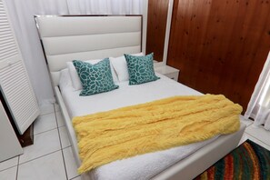 Een bed met een wit hoofdeinde, twee gestreepte kussens en een gele plaid.
