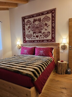 Une chambre avec une tête de lit en bois, une grande tapisserie ornée de motifs naturels et de cœurs, deux lampes fixées au mur, une couverture de lit rouge et un petit tabouret en bois décoré d’une fleur.