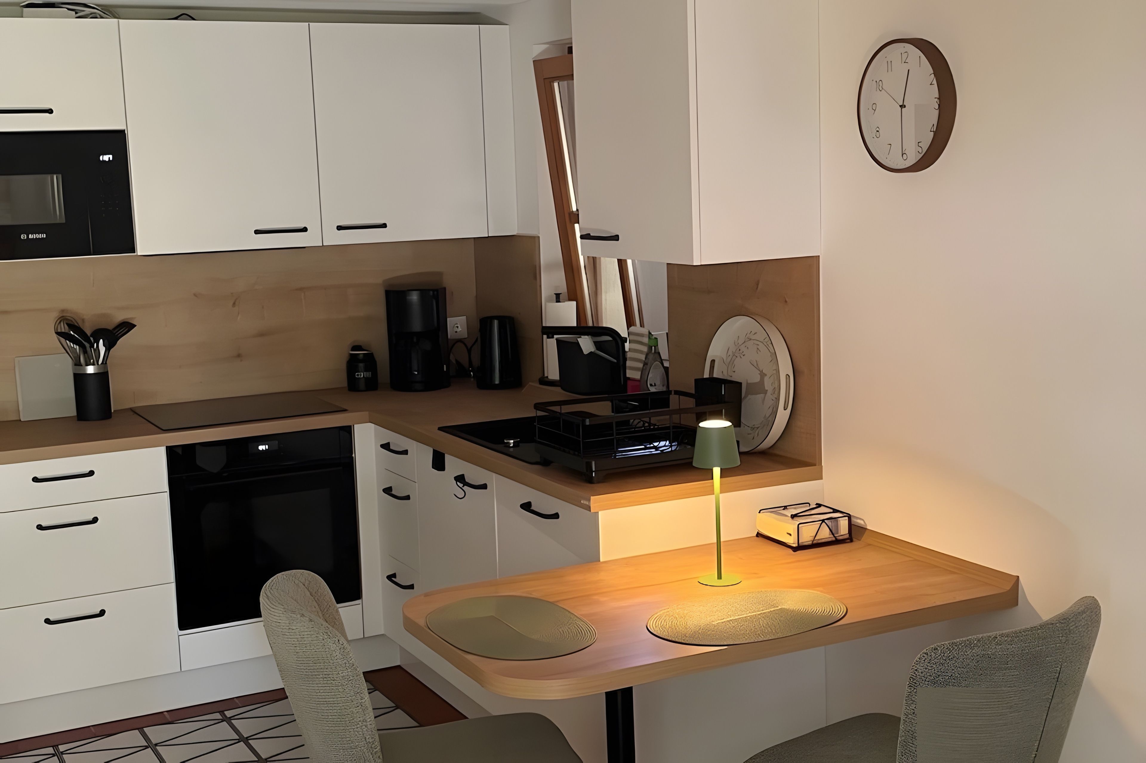 Une cuisine de taille réduite avec des meubles blancs, une cuisinière noire, une machine à café et une horloge au mur.