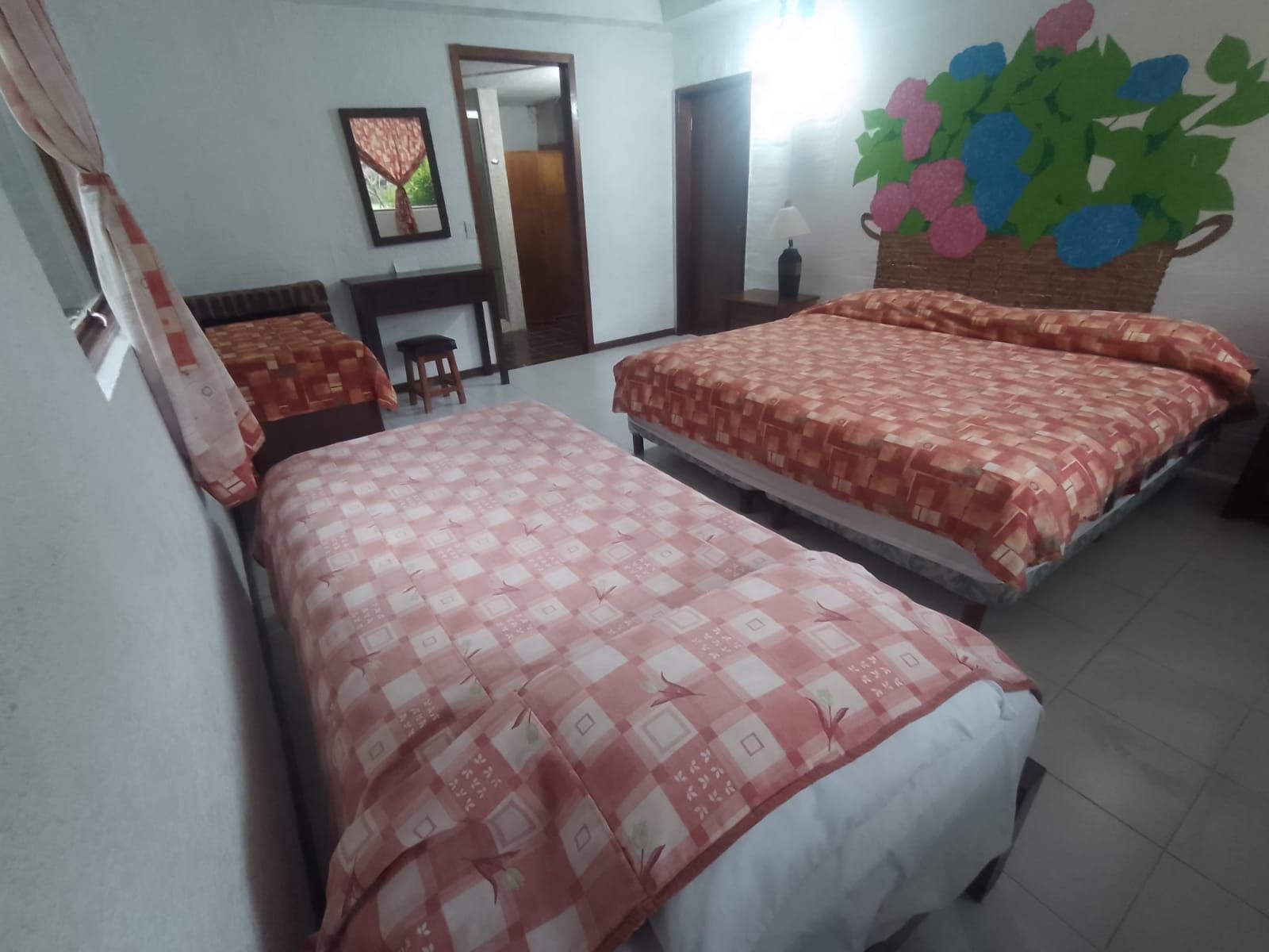 Quarto de hotel com duas camas, uma escrivaninha, uma cadeira, um espelho e um objeto decorativo na parede.