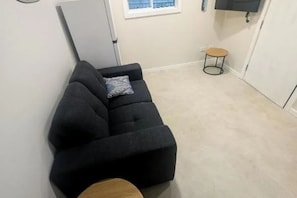 A compact living space with a dark-colored sofa, a small round wooden stool, and a television mounted on the wall. - Fresh & Cosy Beachside Escape (Curl Curl)