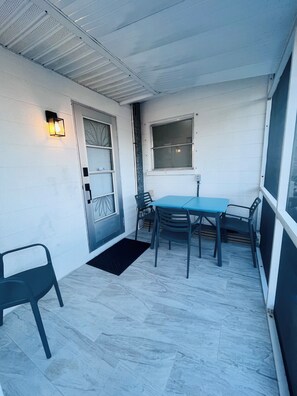 Ein überdachter Patio-Bereich mit Tisch und Stühlen, eine Tür mit Fenster sowie eine wandmontierte Leuchte.