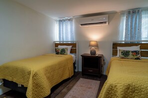 Una habitación con dos camas, una mesita de noche, un aire acondicionado y una ventana con cortinas.