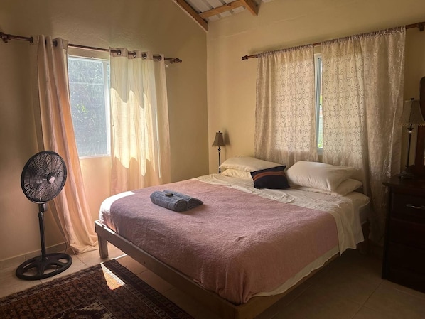 A bedroom with a bed, a standing fan, and a window with curtains. - Casa Baldoria Tropical Escape (Cabrera)