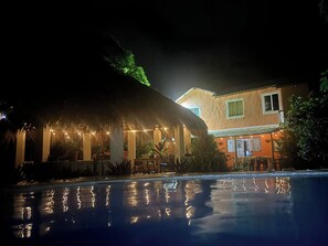 A nighttime view of a resort with a pool, a thatched-roof structure, and a building with windows. - Casa Baldoria Tropical Escape (Cabrera)
