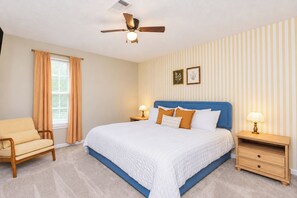 Une chambre à coucher comprenant un lit, une chaise, une table de chevet, une lampe, un ventilateur de plafond et une fenêtre avec des rideaux.