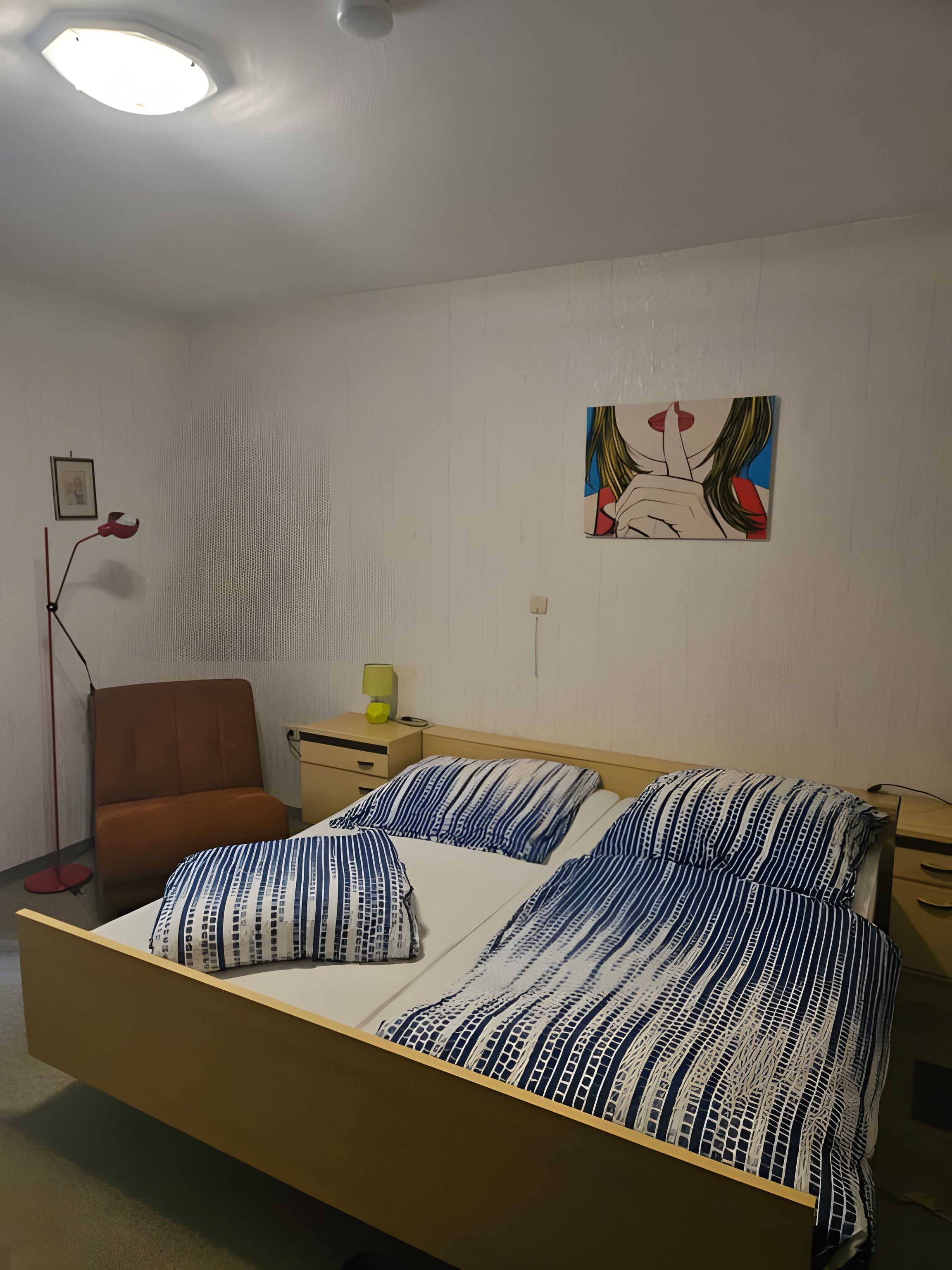 A bedroom with a bed, a chair, a bedside table, a lamp, and a wall with a painting.
