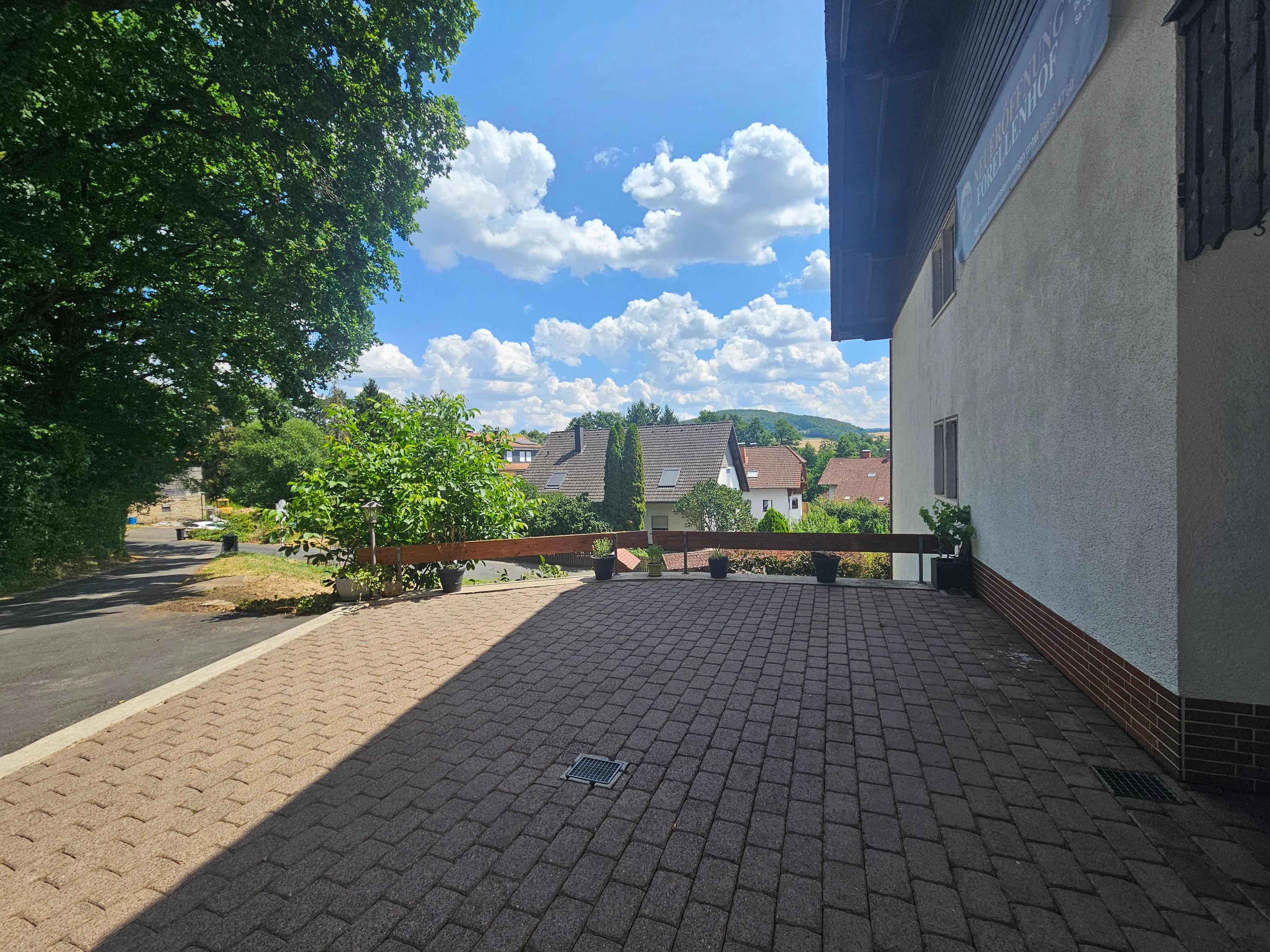 Un espacio al aire libre con suelo pavimentado, cerca de madera, plantas en macetas y vistas a casas y colinas al fondo.