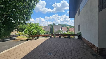 Un espacio al aire libre con suelo pavimentado, cerca de madera, plantas en macetas y vistas a casas y colinas al fondo.