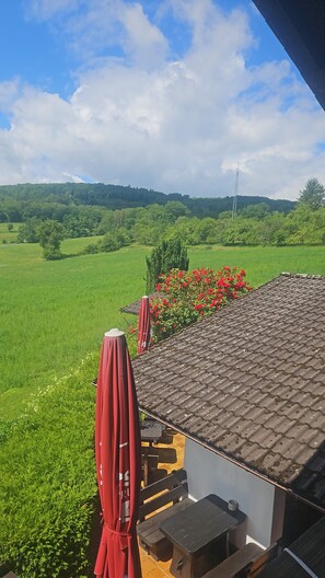 Une terrasse avec vue sur un champ verdoyant et une colline, un parasol rouge et une table avec des chaises.